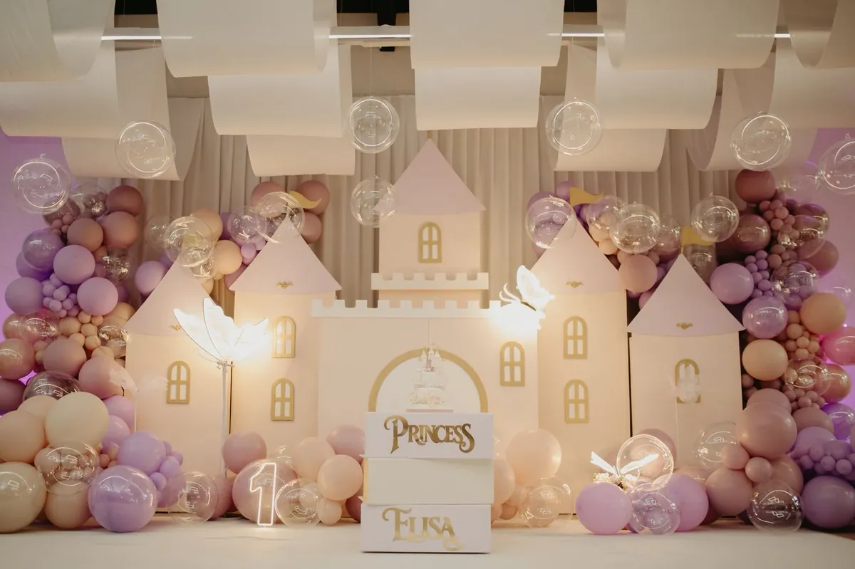 Cenário de castelo cor de rosa e creme, com arcos de balões lilás e o letreiro "Princess Elisa" — festa de 1 ano no 3:16